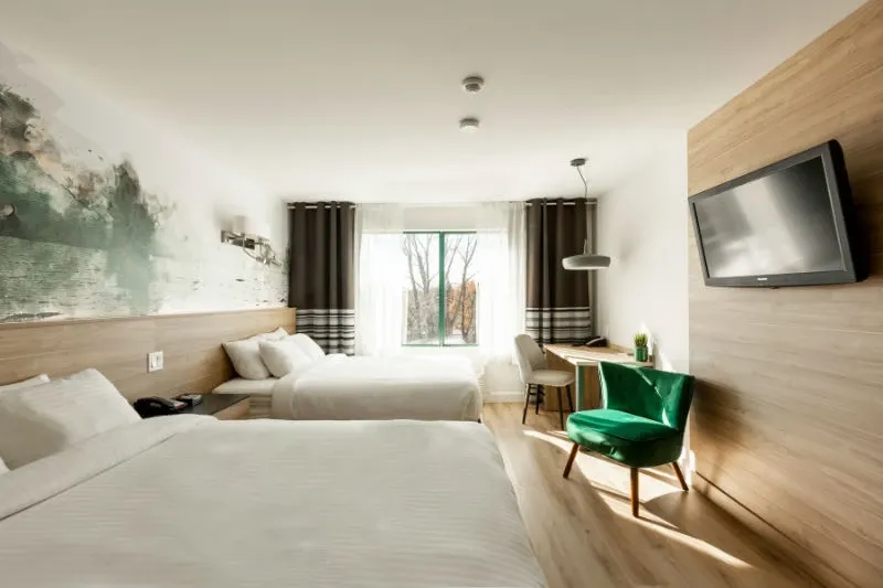 A beautifully lit room at Hôtel Château Joliette, categorized under Wedding Venues, showcasing the elegant accommodations available for weddings. The image features two neatly arranged double beds with crisp white linens, ideal for guests. A large window allows natural light to fill the room, highlighting the modern interior décor, including a unique abstract wall mural. To the right, a flat-screen TV is mounted on the wooden paneled wall above a vibrant green accent chair, adding a touch of luxury. Beneath the window, there is a chic desk setup, perfect for wedding planning or guest use.