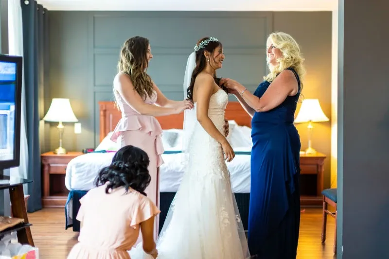 In a beautifully decorated suite at Hôtel Château Joliette, a popular wedding venue in Canada, a bride is surrounded by her bridal party as they assist with her wedding gown. The bride, wearing a stunning white lace dress and a delicate veil adorned with a tiara, stands happily while two women in elegant dresses help with final adjustments. Another woman kneels, arranging the gown's hem. The soft lighting and elegant furnishings create a warm, inviting atmosphere, emphasizing the venue's suitability for a memorable wedding experience.