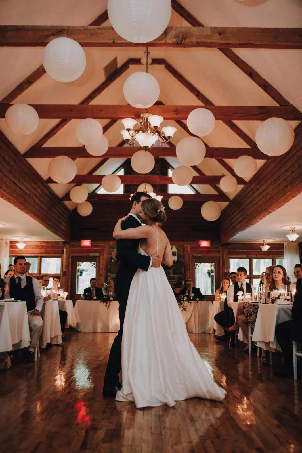 A bride and groom share their first dance in a rustic, elegant setting at Auberge des Gallant, a wedding venue in Canada. The wooden beams of the ceiling are adorned with round paper lanterns, creating a warm, intimate atmosphere. Guests sit at tables decorated with white linens and glowing candles, attentively watching the couple. The bride wears a flowing white gown, while the groom is dressed in a classic dark suit. The image captures a tender moment on the wooden floor, highlighting the romantic and inviting ambiance of this wedding venue.