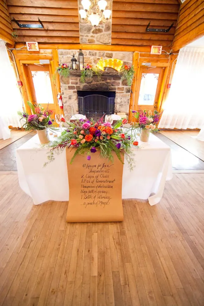 The image showcases a beautifully decorated interior at the Auberge des Gallant, a distinguished choice for wedding venues, part of the Mariage Elegant listings in Canada. The scene features a rustic setting with a prominent stone fireplace adorned with candles and lush greenery. In the foreground, there is an elegantly arranged table with vibrant floral centerpieces, including roses and wildflowers, adding a burst of color to the room. In front of the table hangs a scroll with calligraphy, enhancing the romantic and intimate atmosphere. The warm wooden beams and natural light filtering through sheer curtains further complement the cozy, inviting ambiance ideal for wedding celebrations.