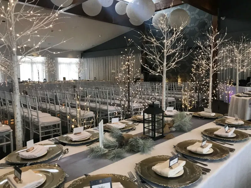 The image shows an elegant indoor wedding setup at Auberge des Gallant, classified under Wedding Venues. The venue features rows of white chairs arranged for a ceremony, with a beautifully decorated banquet table in the foreground. The table is adorned with pine branches, lanterns, and white cotton napkins neatly placed on silver chargers. White birch-like trees with twinkling lights add a winter wonderland atmosphere to the setting, while soft, diffused daylight fills the room through large windows.