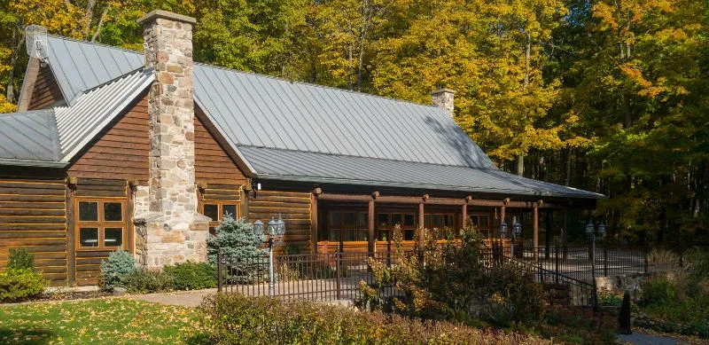 The image showcases the Auberge des Gallant, categorized as a wedding venue, surrounded by lush greenery and vibrant autumn foliage in Canada. The venue features a rustic log building with a stone chimney, adding to its charming and inviting atmosphere. A well-maintained garden with paved walkways leads visitors to the entrance, and the outdoor setting is perfect for picturesque wedding ceremonies or receptions. Ideal for couples seeking a serene and picturesque backdrop for their special day, this location embodies elegance and natural beauty.