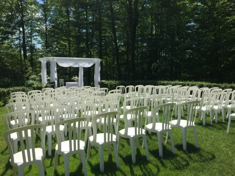 The image showcases an outdoor wedding ceremony setup at Auberge des Gallant, a wedding venue listed on Mariage Elegant in Canada. Rows of white chairs are neatly arranged on a lush green lawn, facing a beautifully decorated white arbor. The arbor is situated against a backdrop of tall trees, creating a serene and picturesque setting perfect for a wedding ceremony. The scene conveys an atmosphere of elegance and tranquility, ideal for couples seeking a natural and charming venue for their special day.