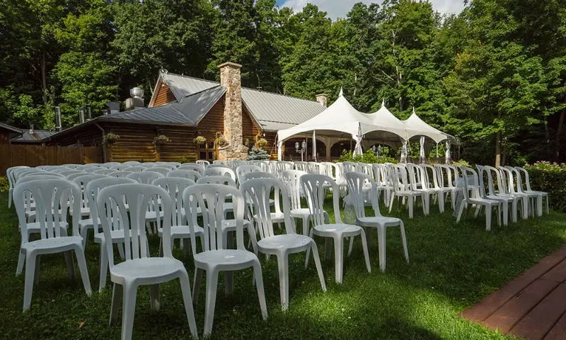 The image showcases the outdoor wedding venue at Auberge des Gallant, nestled within lush greenery. Rows of white chairs are neatly arranged on a well-manicured lawn, facing a rustic building with a stone chimney, ideal for hosting wedding ceremonies. A large white canopy tent is set up nearby, offering covered seating options. Surrounded by a dense forest backdrop, the venue creates a picturesque and intimate setting for wedding celebrations.