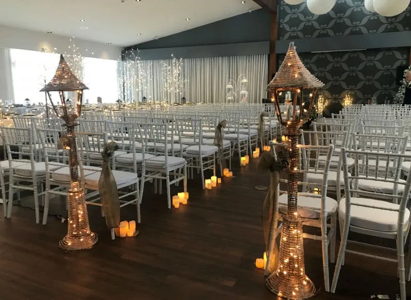 The image shows a beautifully arranged indoor wedding setting at Auberge des Gallant, a wedding venue in Canada. Rows of white chairs are neatly aligned, leading to the altar at the front, creating an elegant aisle. The aisle is adorned with warm, glowing faux candles placed along the floor, adding a romantic ambiance. On either side of the aisle, there are intricately designed woven lanterns with twinkling lights, enhancing the venue's sophisticated atmosphere. The background features softly lit drapes and delicate decorative elements, perfect for a memorable wedding ceremony.