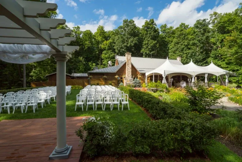 The image shows an outdoor wedding venue at Auberge des Gallant, listed under the Wedding Venues category for Mariage Elegant, Canada. The setting features a rustic building surrounded by lush greenery and mature trees. White chairs are neatly arranged on the grass, facing a decorative pergola, creating a serene and elegant ceremony space. In the background, white tents provide additional covered areas, blending seamlessly with the natural surroundings. The atmosphere is tranquil, making it a picturesque choice for a wedding celebration.