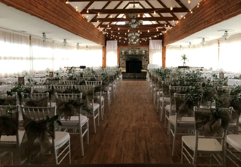 The image showcases the interior of Auberge des Gallant, a wedding venue featured on Mariage Elegant in Canada. The venue is elegantly set up for a wedding ceremony, with rows of white chiavari chairs arranged neatly on either side of a wooden aisle. Each chair is adorned with rustic burlap bows and greenery, adding a natural touch. Overhead, soft string lights create a warm and inviting ambiance. At the end of the aisle, there is a charming stone fireplace, enhancing the cozy atmosphere. The high wooden beams and draped white curtains complete the sophisticated and romantic setting.