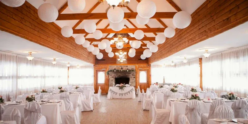 The image showcases a beautifully decorated wedding venue at Auberge des Gallant, categorized under Wedding Venues for Mariage Elegant in Canada. The spacious hall features rustic wooden beams and is adorned with elegant white paper lanterns hanging from the ceiling. The room is filled with round tables draped in white linens, each set with delicate tableware and floral centerpieces, creating a sophisticated and inviting atmosphere. In the background, a charming stone fireplace adds a cozy touch, completing the perfect setting for a memorable wedding celebration.