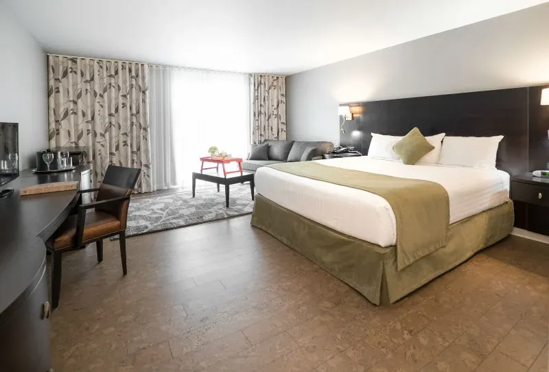 The image showcases a tastefully decorated guest room at Auberge des Gallant, a recognized wedding venue in Canada. The room features a large, neatly made bed with green accents and pillows, complementing the calm, neutral tones of the space. To the left, there is a wooden desk paired with a comfortable chair, ideal for wedding planning activities. Natural light floods the room through sheer curtains, enhancing the ambiance. In the corner, a cozy sitting area with a sofa and coffee table provides additional comfort, making it a welcoming space for wedding guests.
