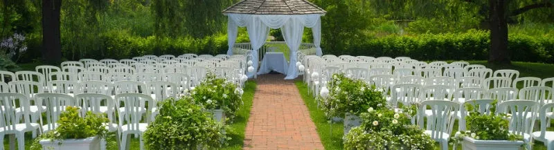 The image showcases the wedding venue at Auberge des Gallant, featuring an outdoor setting arranged for a ceremony. A charming gazebo draped with elegant white fabric serves as the focal point, complemented by lush greenery. Rows of white chairs are neatly aligned on either side of a brick path, creating an inviting aisle. The surrounding natural scenery enhances the romantic atmosphere, making it an ideal location for weddings.