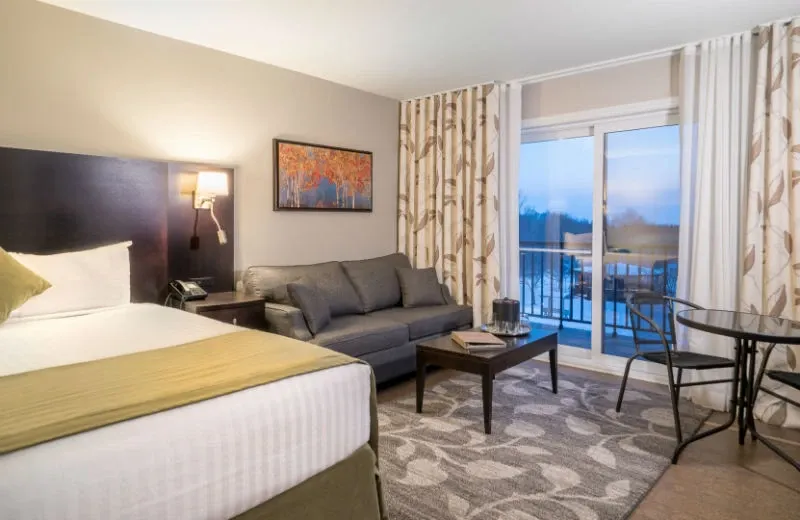 The image shows a cozy guest room at Auberge des Gallant, a distinguished wedding venue in Canada. The room features a comfortable bed with a neatly arranged olive-green pillow and a soft, white duvet. Adjacent to the bed is a sitting area with a stylish gray sofa and a modern coffee table, complemented by a piece of artwork depicting autumn leaves on the wall. Large windows with nature-inspired curtains lead to a balcony that offers a view of the serene outdoors, ideal for moments of relaxation during wedding celebrations. The space is well-lit with a blend of natural and ambient lighting, creating an inviting atmosphere for wedding guests.