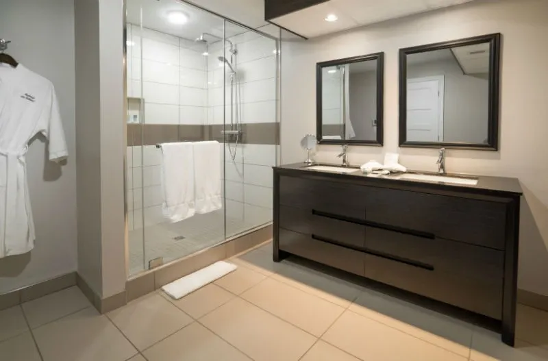 The image showcases a luxurious bathroom at Auberge des Gallant, a wedding venue in Canada. The bathroom features a modern design with a spacious glass shower enclosure and sleek tiling. A dark wood vanity with two mirrors and dual sinks is visible, highlighting the elegance suitable for wedding preparations. A white robe labeled with the venue's name is hanging on the wall, adding a touch of sophistication. This setting perfectly complements the upscale experience offered at this wedding venue.