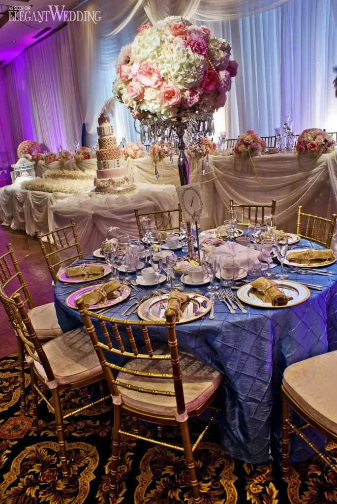 The image showcases an elegantly set wedding reception table at Palace Centre de Congrès, categorized under Wedding Venues for Mariage Elegant, Canada. The table is adorned with a luxurious blue tablecloth and surrounded by gold chiavari chairs. Place settings are meticulously arranged with fine china, glassware, and golden napkin rings. A stunning floral centerpiece, featuring white and pink blooms, complements the intricate decor. In the background, a lavishly decorated multi-tier wedding cake and additional floral arrangements enhance the opulent atmosphere, set against a backdrop of soft, draped fabric with ambient purple lighting.