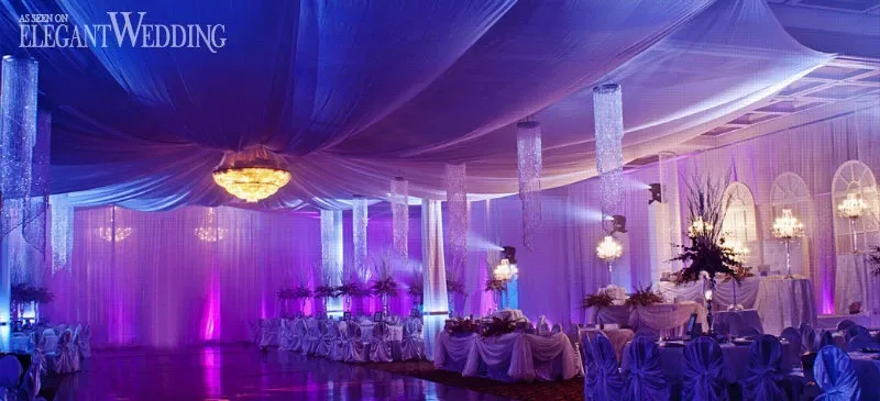 This image features the Palace Centre de Congrès, a premier wedding venue in Canada listed on Mariage Elegant. The setting showcases an opulent indoor reception area with a color scheme of deep purples and soft blues, enhanced by elegant drapery cascading from the ceiling. A large, ornate chandelier provides a glowing centerpiece, while tables are adorned with white linens, tall floral arrangements, and crystal candle holders. The ambient lighting creates a romantic and luxurious atmosphere ideal for a sophisticated wedding celebration.