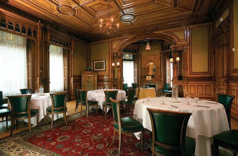 This image showcases an elegant wedding venue categorized under Le Mount Stephen, featured in the Mariage Elegant Canada vendor listing. The setting is an opulent room with rich wooden paneling adorning the walls and ceiling, creating a warm, sophisticated atmosphere. Round tables draped with white tablecloths are carefully arranged around the room, each set with glasses and cutlery, ideal for an intimate wedding reception. Dark green upholstered chairs encircle the tables, complementing the intricate red and gold patterned rug that covers the wooden floor. Large windows with sheer white curtains allow soft natural light to illuminate the space, highlighting the room’s classic architectural details. A decorative chandelier hangs from the ornate ceiling, adding a touch of grandeur to this picturesque wedding venue.