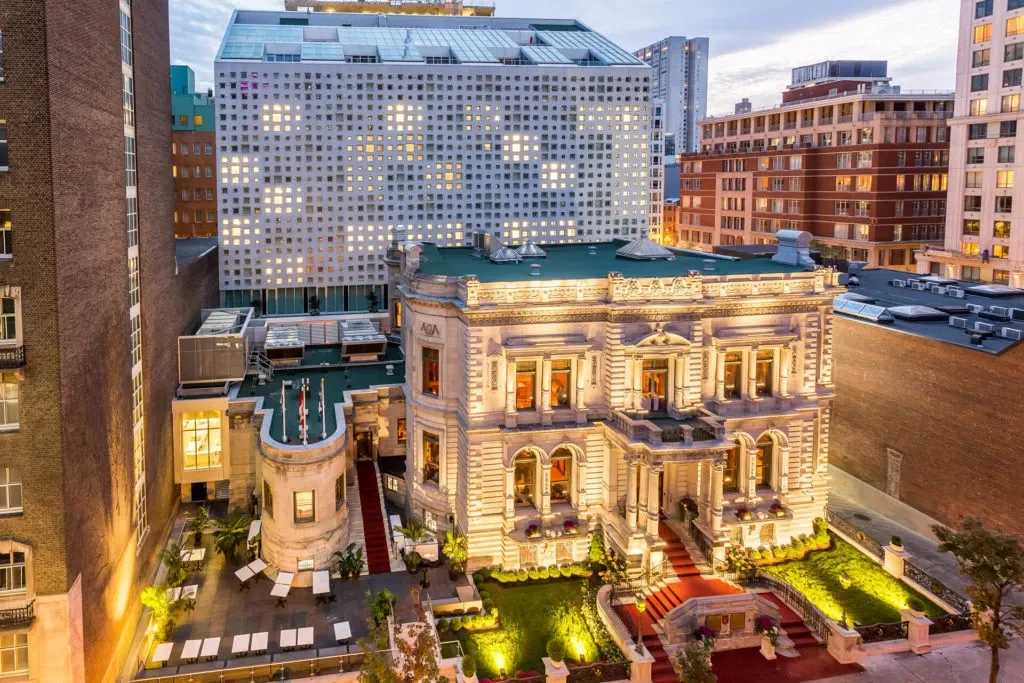 Le Mount Stephen, a premier wedding venue in Canada, is captured in this image showcasing its elegant architecture and grand exterior. The historic building is beautifully illuminated, highlighting its intricate details in the evening light. Surrounded by modern city buildings, the venue stands out with its luxurious charm and well-maintained gardens. The red carpet entrance adds a touch of sophistication, making it a perfect choice for couples seeking a stylish wedding setting.