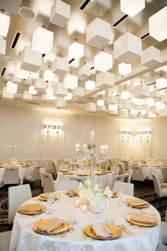 This image showcases an elegant wedding venue at Le Mount Stephen, classified under Wedding Venues on Mariage Elegant, Canada. The venue features a beautifully decorated banquet hall with stylish modern decor. The ceiling is adorned with contemporary white cube light fixtures, creating a sophisticated ambiance. Round tables are elegantly set with white tablecloths, gold charger plates, crystal glassware, and delicate floral centerpieces, exuding luxury and refinement. The soft lighting and pristine setting make it an ideal location for a memorable wedding celebration.