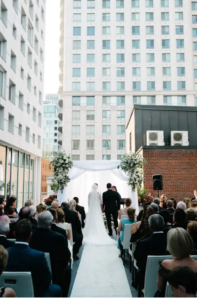 The image showcases an elegant outdoor wedding ceremony at Le Mount Stephen, a premier wedding venue in Canada. The scene is set between modern high-rise buildings, creating a sophisticated urban backdrop. A bride in a flowing white gown and a groom stand before a beautifully adorned archway, decorated with lush white and green floral arrangements. Guests are seated on either side of the aisle, attentively watching the couple. The atmosphere is intimate and stylish, capturing the essence of a chic city wedding.