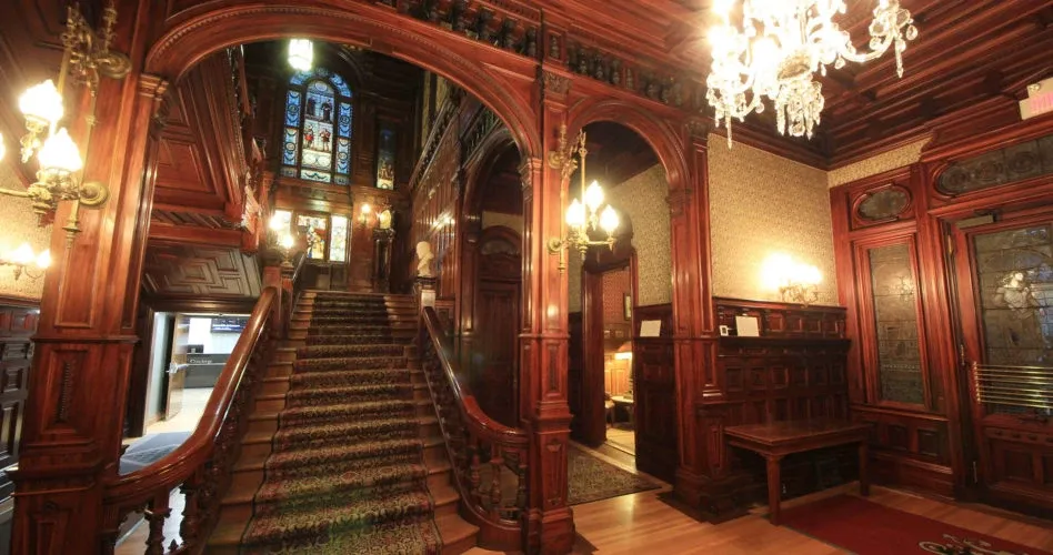 The image showcases the opulent interior of Le Mount Stephen, a distinguished wedding venue in Canada. The grand staircase is adorned with an intricate carpet and leads to a striking stained glass window, adding a touch of elegance and timeless charm. The richly detailed wood paneling and ornate chandeliers illuminate the entrance, creating a warm and inviting atmosphere, perfect for a sophisticated wedding celebration.