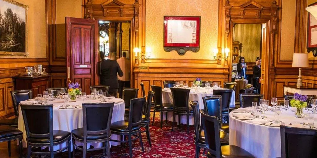 The image showcases an elegant wedding venue at Le Mount Stephen, categorized under Wedding Venues for Mariage Elegant in Canada. The room features ornate wood paneling and sophisticated decor with round tables set for a formal event, each adorned with pristine white tablecloths and vibrant floral centerpieces. Luxurious black chairs complement the classic interior, while refined artwork and warm lighting enhance the upscale ambiance, providing an ideal setting for a stylish wedding celebration.