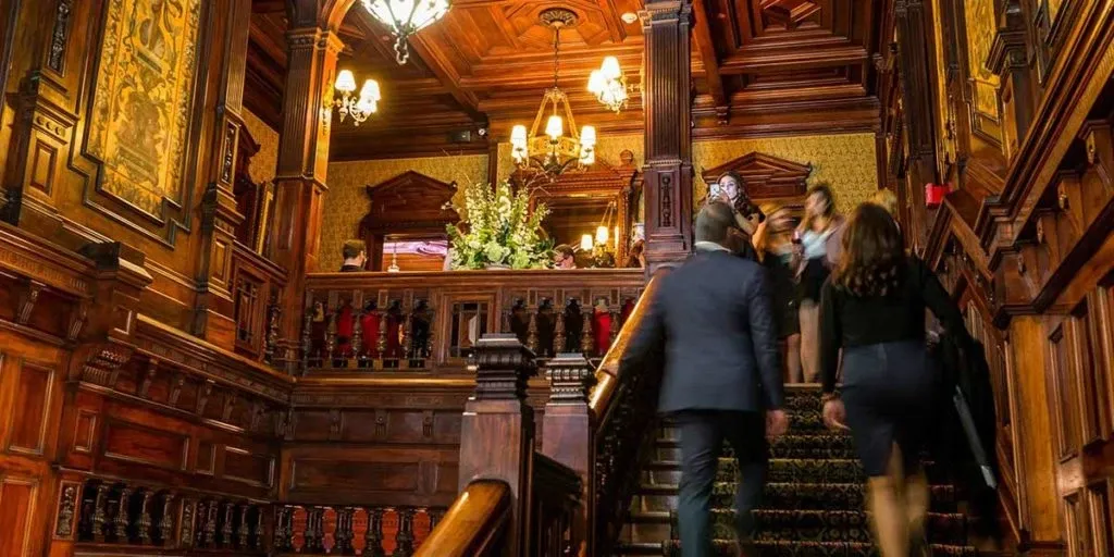 The image shows a luxurious interior of the wedding venue Le Mount Stephen, categorized under Wedding Venues, at Mariage Elegant, Canada. The scene captures an ornate wooden staircase with intricate carvings, leading to an upper level. Elegant chandeliers hang from the richly detailed coffered ceiling, casting warm light throughout the space. Flower arrangements adorn the landing, adding a touch of natural beauty. Guests in formal attire are seen ascending the stairs, highlighting the venue’s suitability for sophisticated wedding events.