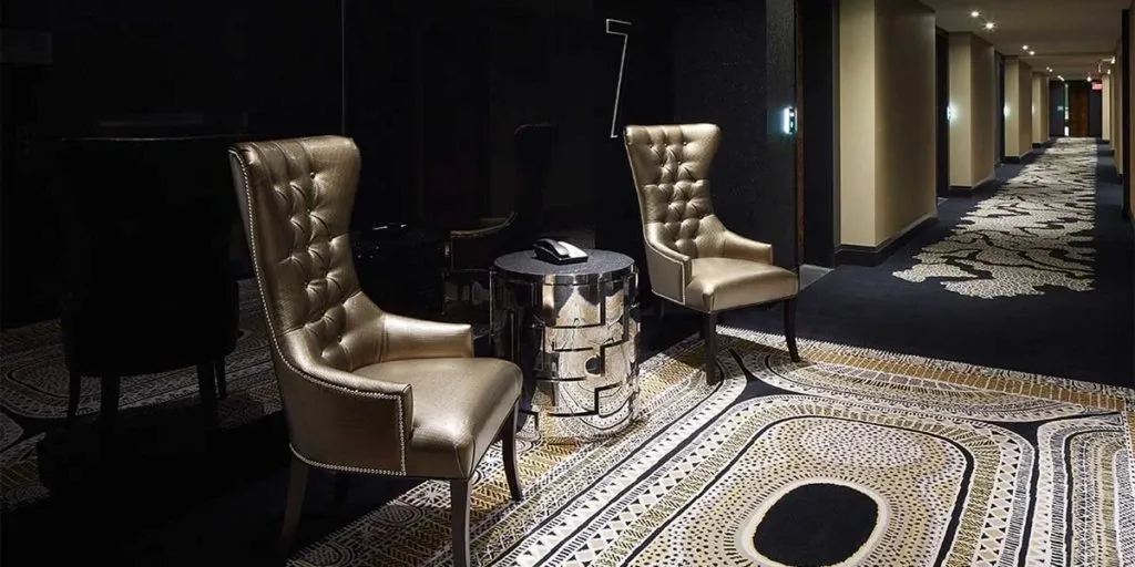 In the image of the elegant wedding venue, Le Mount Stephen, categorized under Wedding Venues on Mariage Elegant, Canada, there is a sophisticated hallway featuring plush, high-backed, metallic-toned chairs arranged around a sleek, reflective side table. The dimly lit corridor boasts a richly patterned carpet that enhances the luxurious ambiance, ideal for intimate wedding gatherings.