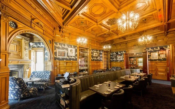 The image showcases the elegant interior of the Le Mount Stephen, a distinguished wedding venue in Canada. The room features stunning wood-paneled walls and a high, intricately designed wooden ceiling adorned with ornate chandeliers that cast a warm glow throughout the space. Several framed paintings and sophisticated decor elements contribute to the luxurious ambiance. Plush seating and meticulously set tables suggest a refined setting suitable for wedding gatherings or receptions, enhancing the allure of booking this venue for an unforgettable event.