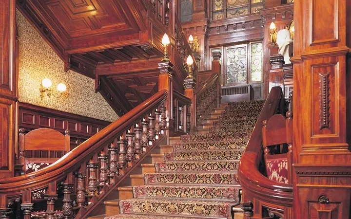 In this image of Le Mount Stephen, a premier wedding venue, the grandeur of the staircase immediately catches the eye. The rich, dark wood of the bannisters and paneling exudes elegance, complemented by ornately carved details. The staircase is carpeted with an intricate floral-patterned runner, adding a touch of luxury. Warm lighting from vintage-style sconces enhances the opulent ambiance. This stunning interior reflects the sophisticated atmosphere ideal for a wedding or special event at Le Mount Stephen in Canada.