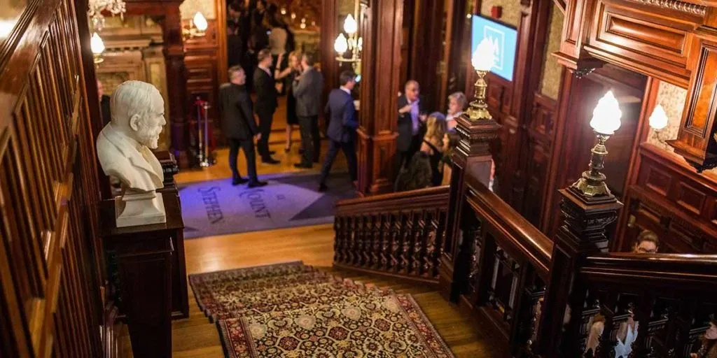 The image showcases the elegant interior of Le Mount Stephen, a premier wedding venue listed on Mariage Elegant, Canada. The scene captures an opulent staircase adorned with intricate woodwork and a richly patterned carpet. A white marble bust is situated prominently, adding a touch of sophistication. Warm lighting from ornate lamps enhances the luxurious ambiance. Several well-dressed guests can be seen mingling near the entrance, suggesting a lively event, possibly a wedding reception. The refined décor and atmosphere highlight Le Mount Stephen as an ideal setting for weddings.