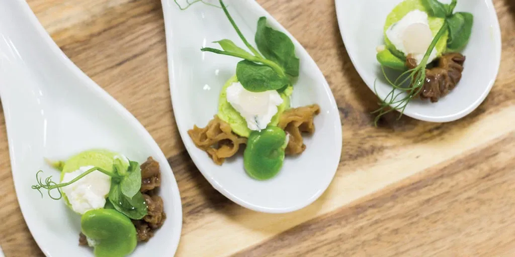 Exquisite hors d'oeuvres elegantly presented on white ceramic spoons are displayed at Le Mount Stephen, showcasing the venue's fine dining options. Each spoon holds a delicate arrangement of fresh green pea puree, crumbled cheese, and garnished with pea shoots, highlighting the attention to detail and gourmet offerings available at this wedding venue. The wooden surface adds a rustic touch, enhancing the sophisticated presentation perfect for a wedding reception, making Le Mount Stephen a distinguished choice in the wedding venues category for Mariage Elegant, Canada.