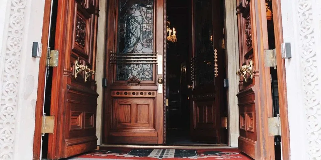 This image showcases the grand entrance of Le Mount Stephen, an elegant wedding venue in Canada. The wooden double doors are intricately carved with ornate designs, reflecting a sophisticated and historical ambiance. A glimpse of a luxurious chandelier is visible inside, enhancing the venue's opulent atmosphere. The red and white patterned carpet adds a touch of refinement, creating a welcoming path for wedding guests. Le Mount Stephen offers a picturesque setting for couples seeking a timeless and elegant backdrop for their special day.