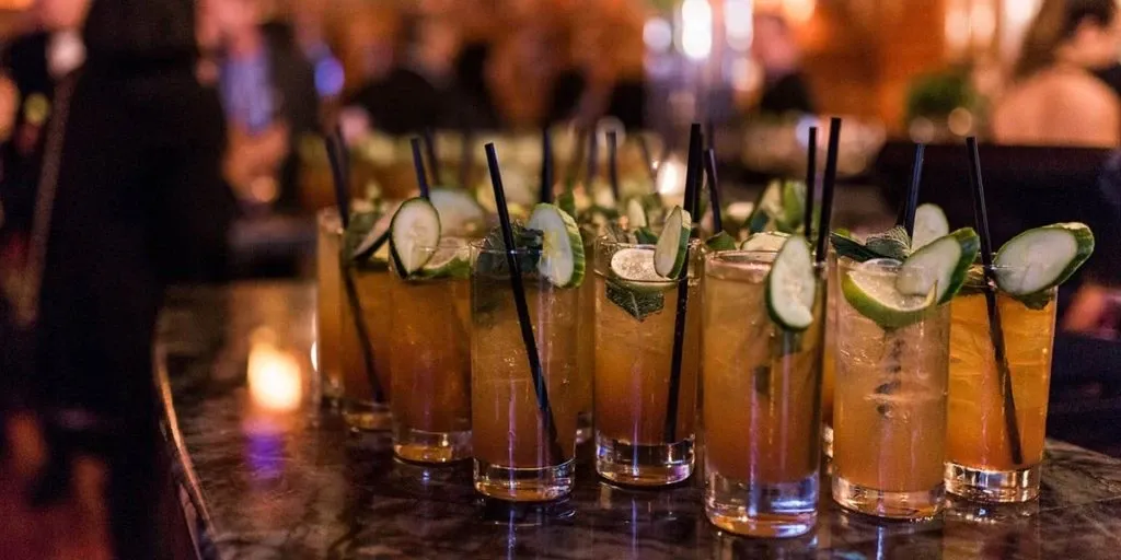 A row of elegant cocktails garnished with cucumber slices and mint leaves is displayed on a polished marble bar top. The setting is dimly lit, creating a warm and inviting ambiance perfect for a wedding reception. This scene captures the sophisticated atmosphere at Le Mount Stephen, showcasing its potential as a stunning wedding venue. Guests in the background engage in conversation, adding to the lively and celebratory mood of the occasion.