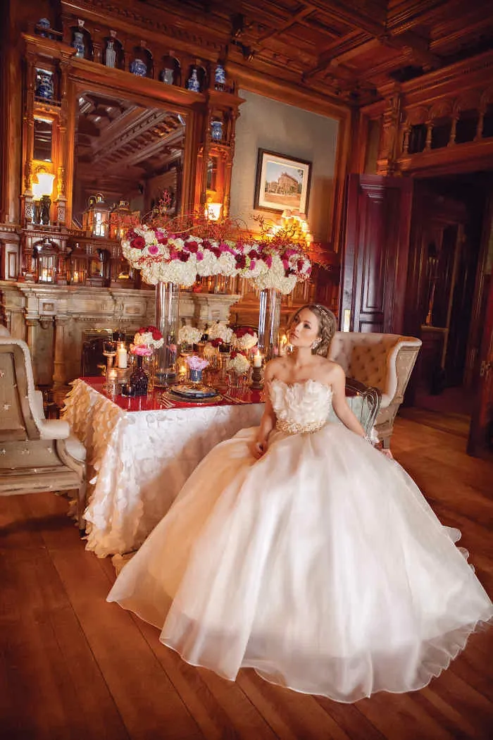 In this image, a bride in an elegant, flowing white gown is seated beside a beautifully decorated table at Le Mount Stephen, highlighting its charm as a premier wedding venue. The table is adorned with an array of vibrant floral arrangements featuring red, pink, and white flowers, adding a romantic touch to the sophisticated setting. The backdrop showcases ornate wood paneling, intricate carvings, and a decorative mirror, contributing to the venue’s luxurious ambiance. Warm lighting and candles create an intimate atmosphere, perfect for weddings hosted by Le Mount Stephen.
