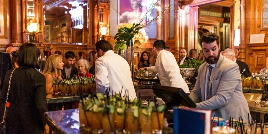 This image showcases the elegant interior of Le Mount Stephen, highlighting its role as a premier wedding venue in Canada. The setting features a lavish bar area with intricately carved wooden details and a large mirror reflecting the warm ambiance. Bartenders in white jackets are preparing drinks, while guests gather around, enjoying cocktails adorned with cucumber slices. The sophisticated atmosphere, combined with the attentive service, makes Le Mount Stephen an ideal choice for couples seeking a luxurious wedding experience.