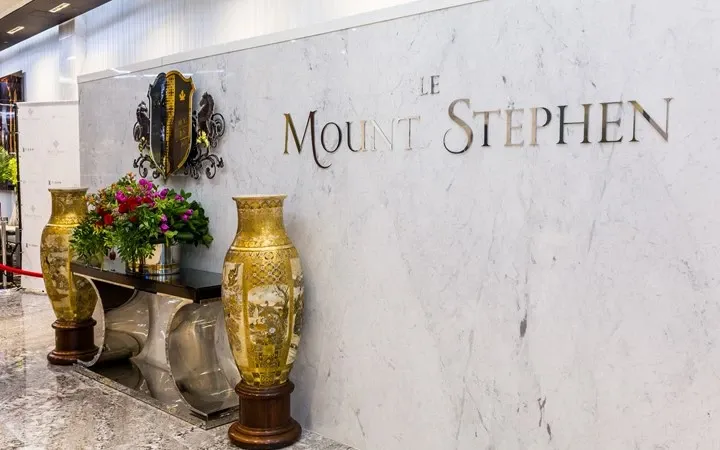 The image features the elegant entrance area of Le Mount Stephen, a premium wedding venue listed under Mariage Elegant, Canada. The scene showcases a luxurious marble wall with the venue's name, 'Le Mount Stephen,' prominently displayed in refined lettering. To the side, ornate, gold-accented vases are positioned on either side of a collection of vibrant floral arrangements, adding to the sophisticated ambiance. The decor highlights the venue's dedication to offering an upscale and stylish setting for weddings and special events.