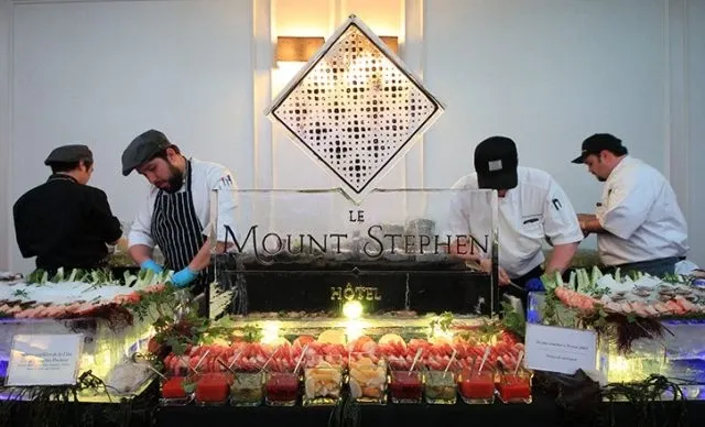 The image showcases a luxurious buffet setup at Le Mount Stephen, a top wedding venue in Canada. In the foreground, a variety of elegantly presented appetizers and hors d'oeuvres are arranged on a glass-topped table adorned with greenery. Three chefs, dressed in professional attire, prepare and arrange the exquisite dishes with precision and care. An ice sculpture prominently featuring the name 'Le Mount Stephen' serves as a stunning centerpiece, encapsulating the venue's sophisticated atmosphere, perfect for hosting memorable wedding celebrations.