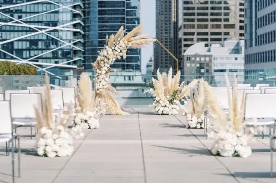 The image shows an elegant outdoor wedding venue setup by Le Mount Stephen, featured in the Wedding Venues category on Mariage Elegant, Canada. The setting includes a beautifully arranged ceremonial aisle on a rooftop with a backdrop of modern city skyscrapers. The aisle is adorned with white chairs on either side and lined with decorative floral arrangements combining pampas grass and white blossoms. At the end of the aisle, a stunning arch made of pampas grass and white flowers creates a focal point for the ceremony space, perfect for a sophisticated urban wedding.