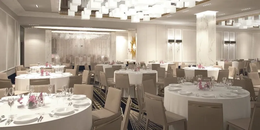 The image shows an elegant wedding venue at Le Mount Stephen, categorized under Wedding Venues. The room is beautifully set up with round tables covered in white tablecloths, surrounded by beige chairs. Each table is adorned with delicate pink floral centerpieces and gleaming glassware, creating a sophisticated atmosphere. The ceiling features modern, square pendant lights, casting a warm glow over the space. The decor is refined, with marble accents and a minimalist aesthetic, ideal for a chic and memorable wedding celebration.