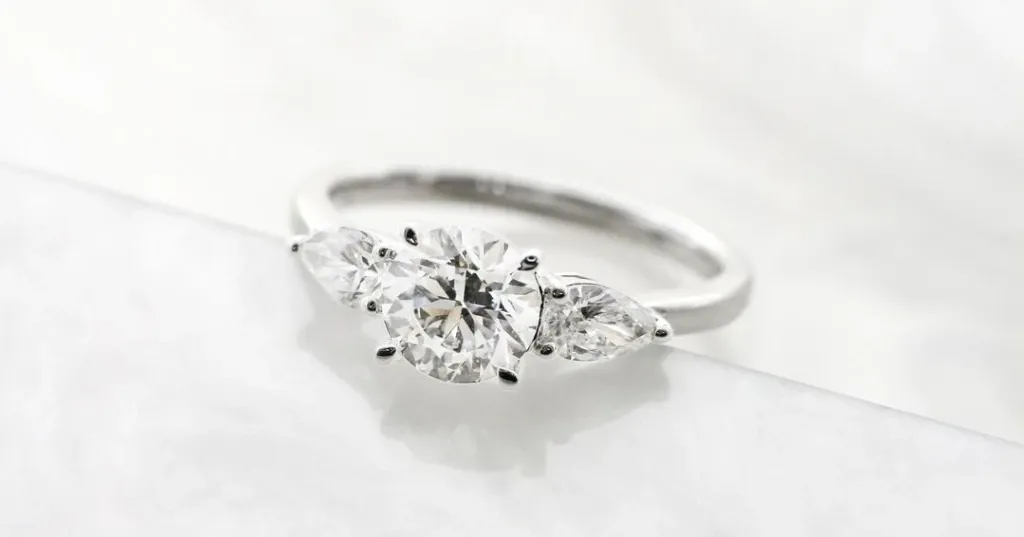 A stunning diamond engagement ring designed by Bijouterie B. Serkos, featured in the Wedding Jewelry category. The ring showcases a brilliant round center diamond flanked by two elegant pear-shaped diamonds, all set in a sleek platinum band. Perfect for couples seeking timeless elegance in their wedding jewelry, this piece exemplifies exquisite craftsmanship suitable for a sophisticated bridal ensemble.