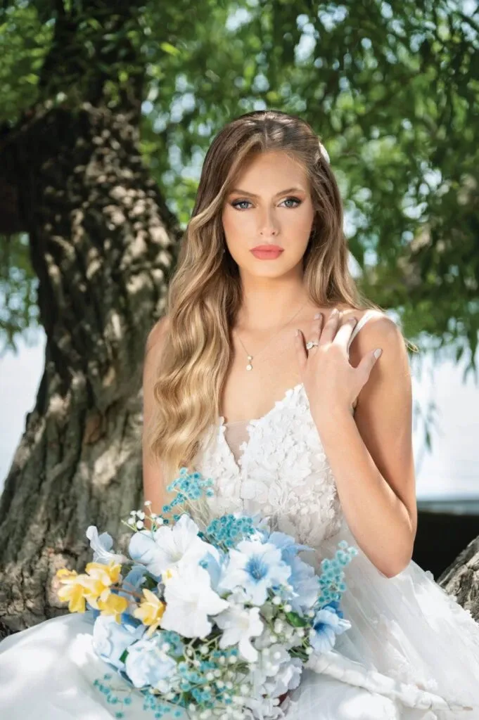 A bride wearing elegant wedding jewelry from Bijouterie B. Serkos, featured under the Wedding Jewelry category, is sitting outdoors. She showcases a delicate necklace and a stunning ring, complementing her intricate white lace wedding gown. The bouquet she holds includes white, blue, and yellow flowers, providing a fresh and vibrant touch. An idyllic, leafy background enhances the romantic setting, ideal for a wedding scene in Canada.