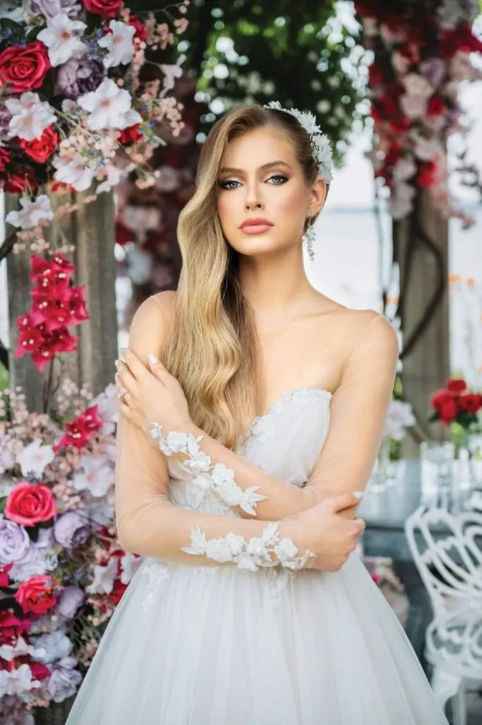 A bride stands elegantly in a stunning wedding dress surrounded by vibrant floral arrangements consisting of red, pink, and white flowers. Her hair cascades in soft waves, adorned with delicate flowers, while she wears exquisite earrings, highlighting her elegant look. This wedding jewelry from Bijouterie B. Serkos, a vendor listed in the Wedding Jewelry category of Mariage Elegant, Canada, adds a touch of sophistication and charm to this beautiful bridal ensemble.