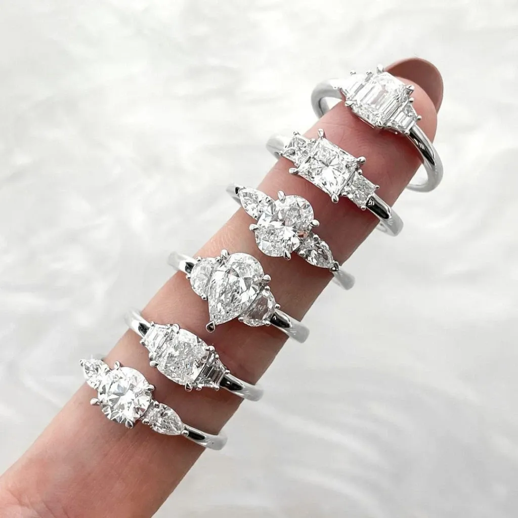 A hand showcases five elegantly designed engagement rings, each featuring sparkling diamonds in various cuts, including oval, cushion, and emerald shapes, set in sleek silver bands. These stunning pieces are from Bijouterie B. Serkos, a vendor specializing in wedding jewelry for the Mariage Elegant website in Canada. The rings rest on a delicate, soft background, emphasizing their brilliance and quality craftsmanship, ideal for any couple planning their special day.