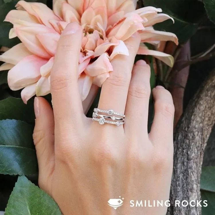 A close-up image of a hand adorned with elegant wedding jewelry from Bijouterie B. Serkos, featured in the wedding planning and vendor listing site, Mariage Elegant in Canada. The hand showcases a beautifully crafted diamond engagement ring and wedding band, both intricately designed with a series of sparkling diamonds. The jewelry complements the hand resting gently against a backdrop of lush green leaves and a blooming pink flower, emphasizing a natural and romantic setting ideal for a wedding.