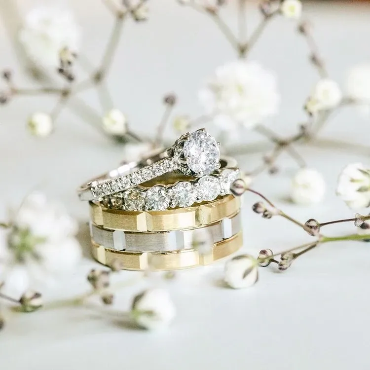 The image features a stunning set of wedding rings designed by Bijouterie B. Serkos, categorized under wedding jewelry. There are three rings: a solitaire engagement ring with a prominent round diamond set atop a band encrusted with smaller diamonds, a matching diamond-studded wedding band, and a contrasting gold and silver band with an elegant geometric pattern. The rings are artistically arranged on a delicate backdrop of white flowers, evoking a sense of elegance and romance suitable for any wedding occasion.