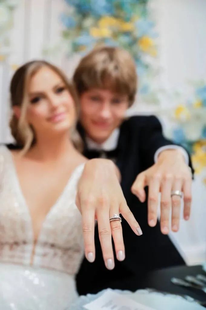 This image features a newlywed couple joyfully displaying their wedding rings, highlighting the exquisite craftsmanship of Bijouterie B. Serkos in the wedding jewelry category. The bride, wearing a beautiful lace wedding gown, and the groom, in a classic black suit, are both smiling warmly. Their hands are prominently shown, emphasizing the elegant rings that symbolize their union. In the background, soft floral decorations in hues of blue and yellow add a romantic and festive touch, setting the perfect backdrop for this matrimonial moment. This image showcases the quality and elegance offered by Bijouterie B. Serkos for discerning couples planning their special day in Canada.