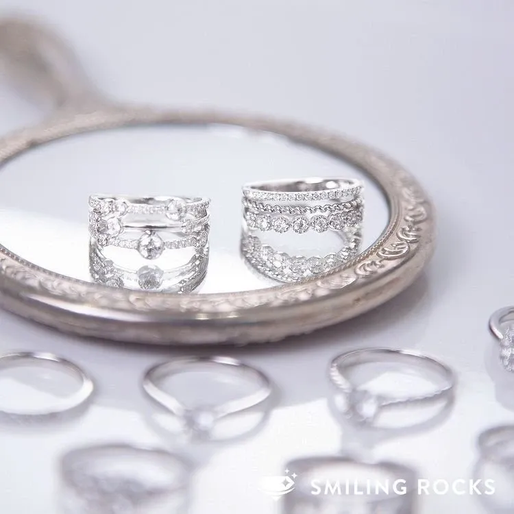The image showcases exquisite wedding jewelry by Bijouterie B. Serkos, beautifully displayed on an ornate, vintage-style hand mirror. Two stunning rings, adorned with sparkling diamonds, are centrally positioned, highlighting their intricate designs and craftsmanship. Surrounding the mirror are additional elegant rings, each reflecting the delicate and luxurious aesthetic perfect for a sophisticated wedding. This image underscores Bijouterie B. Serkos' expertise in crafting exceptional jewelry pieces, ideal for brides seeking timeless elegance for their special day in Canada.