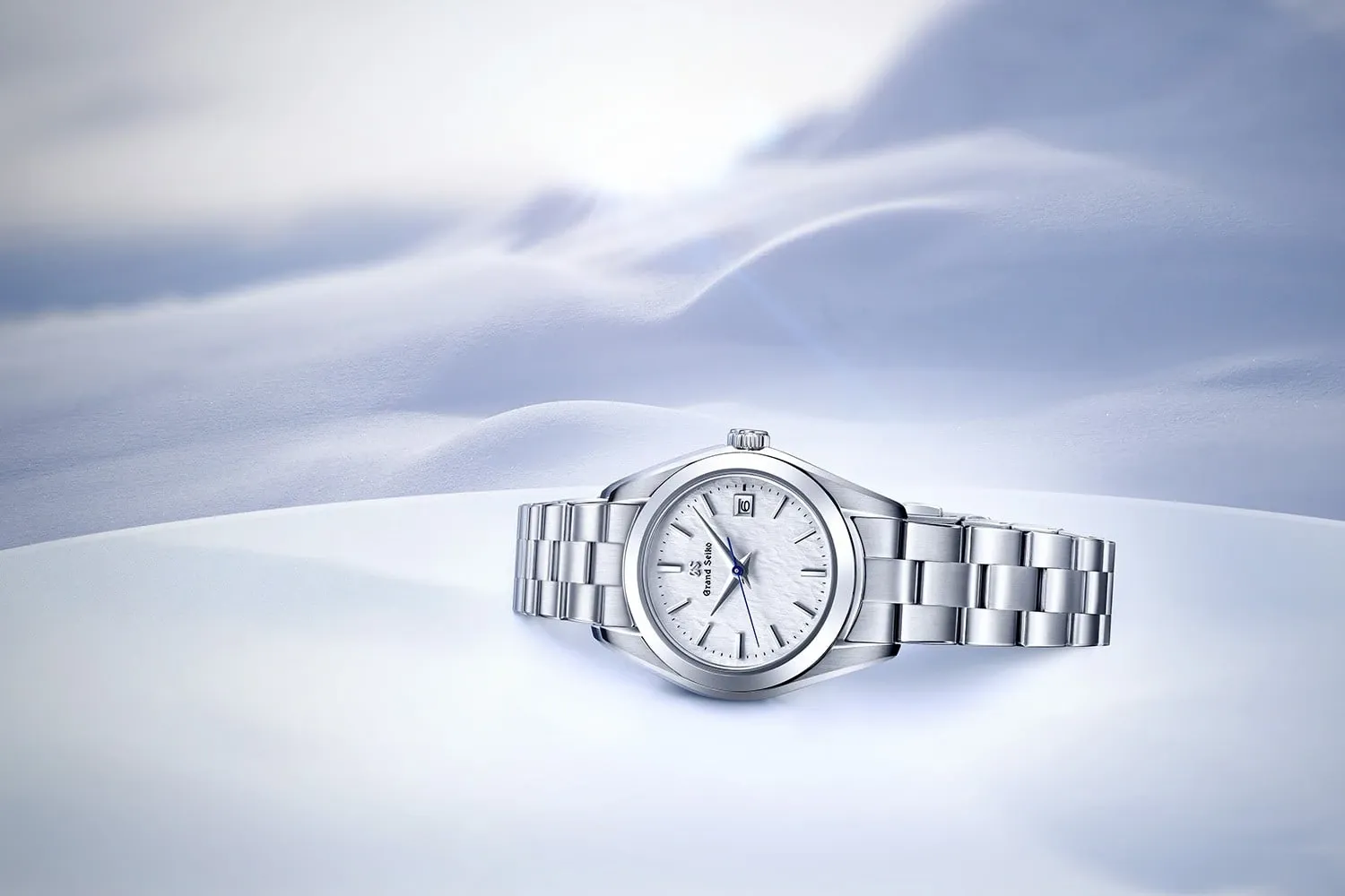 The image features an elegant silver wristwatch displayed on a smooth, white surface, set against a softly blurred background that resembles gentle snowdrifts. This exquisite piece by Bijouterie B. Serkos is ideal for wedding jewelry, adding a touch of sophistication and timeless elegance to any wedding ensemble. The watch's sleek design and polished finish are highlighted, showcasing its quality craftsmanship and luxurious aesthetic, perfect for complementing bridal attire.