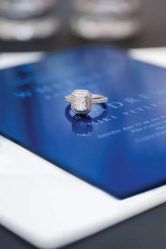 A stunning engagement ring from Bijouterie B. Serkos, categorized under wedding jewelry, is elegantly showcased. The ring features a dazzling rectangular center stone surrounded by a halo of smaller diamonds, set on a band encrusted with additional diamonds. The jewelry piece is placed atop a deep blue wedding invitation, suggesting the sophisticated and luxurious theme of a wedding. The meticulous craftsmanship and sparkle of the ring highlight its suitability for a special occasion, making it an ideal choice for those seeking elegance in their wedding jewelry selection.