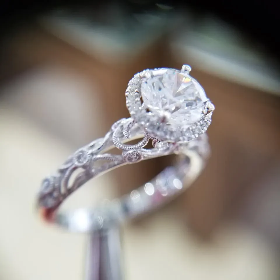 This image showcases an exquisite engagement ring from Bijouterie B. Serkos, featured under the Wedding Jewelry category. The ring boasts a dazzling, large round-cut diamond set in a delicate, intricate band adorned with smaller diamonds and elaborate detailing, highlighting the exquisite craftsmanship. The soft focus background brings attention to the ring's elegant design, ideal for brides seeking timeless sophistication in their wedding jewelry.