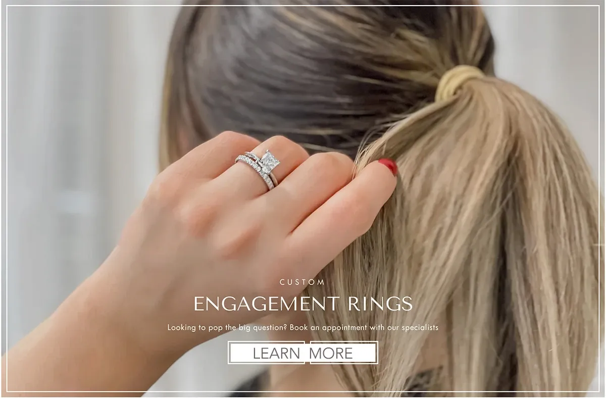 A woman is showcasing an elegant engagement ring set from Bijouterie La Voûte, a vendor in the wedding jewelry category for Mariage Elegant, Canada. The focus is on her hand, highlighting a beautifully designed diamond ring, featuring a prominent square-cut stone accompanied by matching bands encrusted with smaller diamonds. Her hair is styled in a simple ponytail, providing a neutral background that accentuates the jewelry. The image text advertises custom engagement rings with an invitation to learn more about booking an appointment with their specialists.